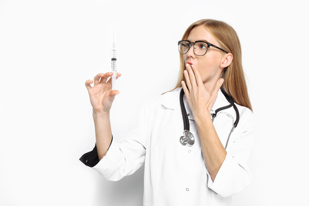 Young scared intern doctor wearing glasses with a stethoscope around his neck, holding a syringe in his hands and looking at him with fear, on a white backgroundの写真素材