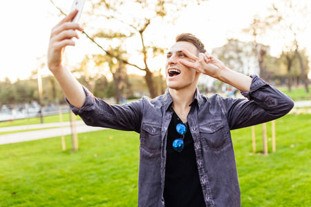 Portrait of a laughing man, wearing sunglasses, standing in a park with a smartphone. He takes...