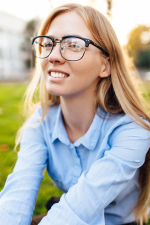 Portrait of a cute young girl in glasses and shirt sitting on the grass in the parkの写真素材