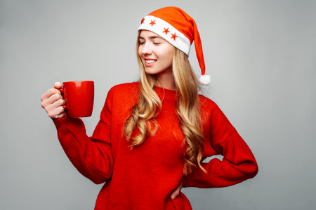 Beautiful woman in a red sweater, in the hat of Santa Claus, with a red mug of coffee, on a gray background. Christmasの写真素材