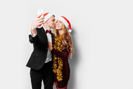 Happy couple of lovers in Santa Claus hats, the guy kisses the girl and makes a photo on the phone, with Christmas gifts in their hands. On a white background.の写真素材