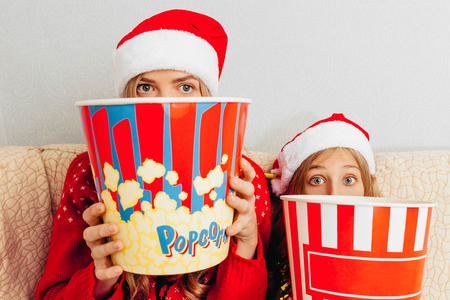 Stunned mother and her little daughter, dressed in Santa Claus hats, are watching movies and eating popcorn, sitting on the couch. Christmas conceptの写真素材