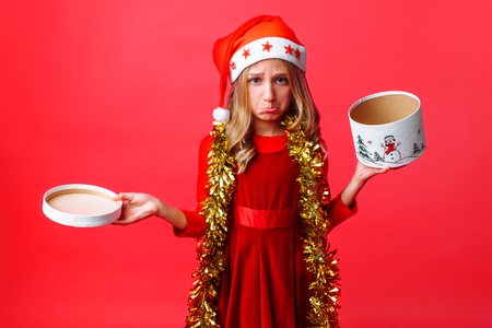 Frustrated teen girl in Santa hat and with tinsel on her neck, with disappointment holding empty box on red backgroundの写真素材