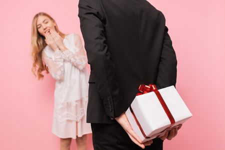 Elegant man in a suit makes a surprise to a woman, gives a bouquet of flowers and a box with a gift, on a pink background, concept of women's dayの写真素材