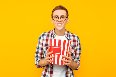 Portrait of a happy man in transparent glasses with a bucket of popcorn, keen on watching a film, in a plaid shirt, isolated on a yellow backgroundの写真素材