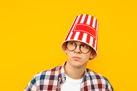 Cheerful man in glasses is happy to watch a movie, with a bucket of popcorn on his head, on a yellow backgroundの写真素材