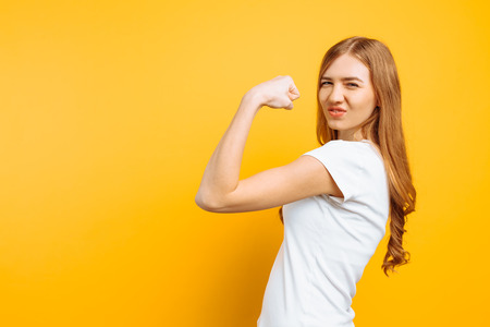 Portrait of happy girl in white t-shirt showing arm muscles on yellow background.