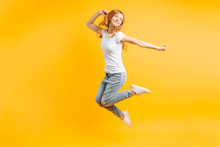 Full length, Portrait of a cheerful enthusiastic girl in a white T-shirt jumping for joy on a yellow backgroundの写真素材