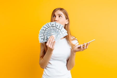 Portrait of an excited woman in white t-shirt showing money bills and holding mobile phone isolated on yellow backgroundの写真素材