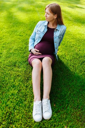 Happy young pregnant woman sitting in the park on the lawn enjoying nature hugging bellyの写真素材