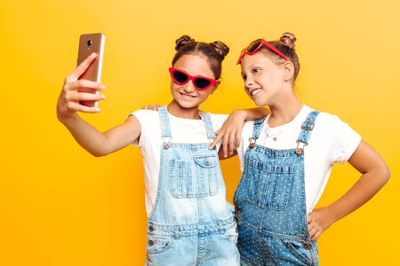 Two teenage girls in sunglasses are resting and taking a selfie on a smartphone on a yellow...