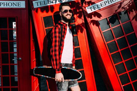 Stylish man with a beard, hipster dressed in a white T-shirt and a plaid shirt, a man holding a skateboard in his hands and posing in front of a red telephone boxの写真素材