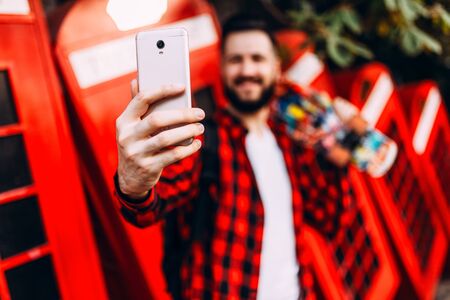 Stylish man with a beard, a hipster dressed in a plaid shirt with a skateboard, takes a selfie on a...