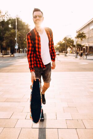 Stylish guy with a beard in sunglasses, walks with a skateboard at sunset.の写真素材