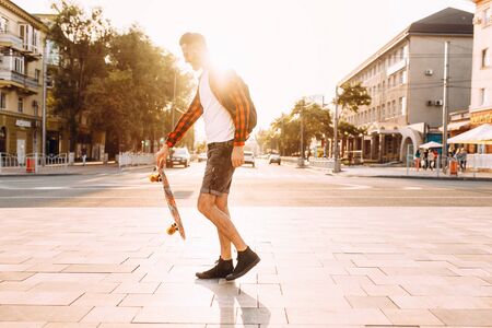 Stylish guy with a beard in wireless headphones walks around the city with a skateboard and listens to musicの写真素材