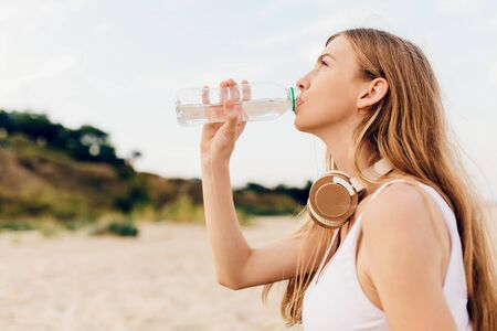 Close-up of tired young girl drinking water from bottle after workout, outdoors at sea