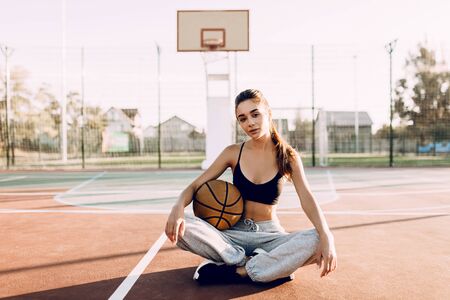 Attractive young athletic girl holding a basketball while sitting in a sports stadium, outdoors. Rest after trainingの写真素材