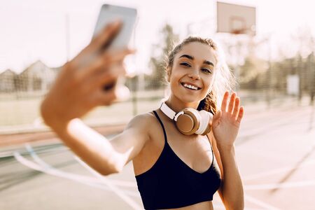 Cheerful athletic woman in sportswear, listening to music with headphones, and taking selfies outdoors after runningの写真素材