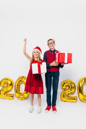 Happy little girl and boy, wearing Santa hat, stylish brother and sister, holding Christmas gifts, and show victory gesture while looking at camera, on white background with gold 2020 figures. The Concept Of The New Year 2020の写真素材