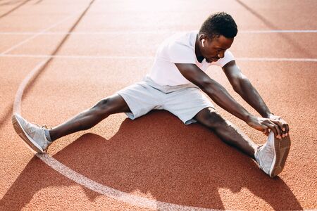 Athletic African-American doing stretching exercises, preparing for a morning workout at the stadium at dawnの写真素材
