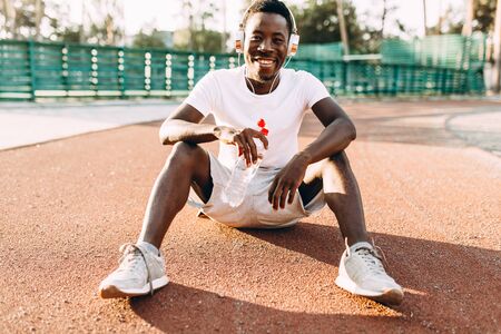 A young athletic African American relaxes after an intense workout. A man rests sitting on the floor at the stadium in the Park, holding a bottle of water,の写真素材