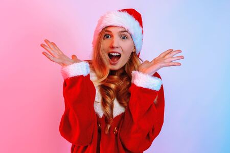 An image of a shocked excited young lady, in a Christmas sweater and Santa hat, standing against a background with red and blue neon lights . Christmas, New Yearの写真素材