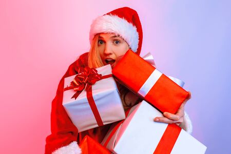 A surprised blonde in a Santa hat looks out from a pile of gifts, stands against a background with a neon lightの写真素材