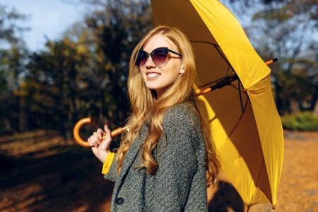 Happy young girl wearing warm coat with yellow umbrella walking in Park with yellow leaves, autumn timeの写真素材