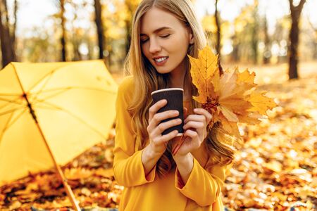 Beautiful happy young girl in autumn Park, with colorful autumn background, sitting on blanket, drinking hot drink, holding autumn leaves in hands, enjoying warm Sunny weatherの写真素材