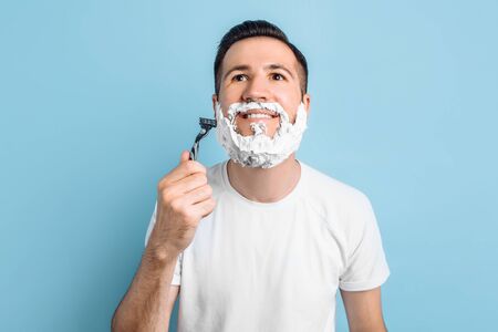 A handsome young man with a beard stands on a blue background with shaving foam on his face looks at the camera and shavesの写真素材
