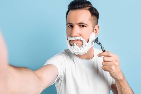 Happy young hipster with a beard, shaves, has foam on his face, holds a razor, looks confidently at...