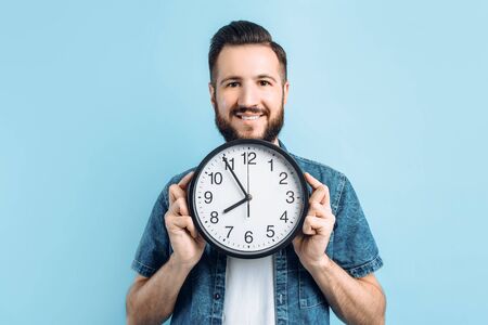 Beautiful stylish happy young man with beard holding round wall clock isolated on blue background