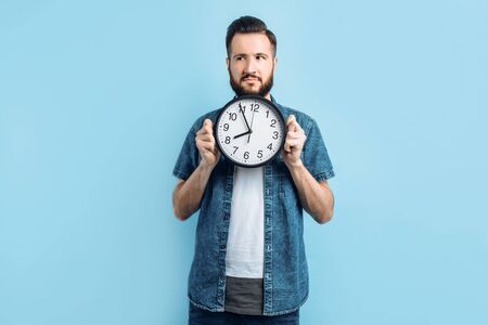 A pensive young man with a beard holds a wall clock, standing on an isolated blue background