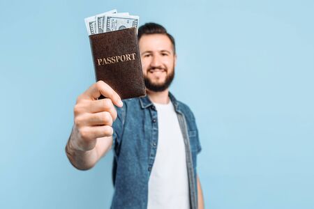 Excited happy young man holding a passport with money posing in isolation on a blue background