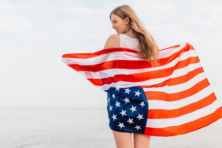 Beautiful Patriotic happy woman with an American flag in her outstretched hands standing on the beachの写真素材