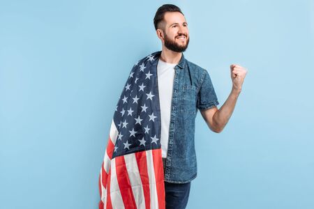 Portrait of a happy young man with a beard , in casual clothes, holding the us flag and demonstrating a gesture of victory and success, on a blue backgroundの写真素材