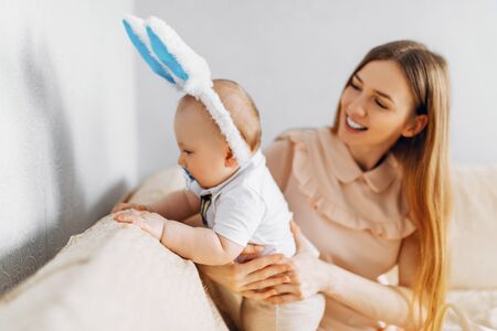 Mom and baby with rabbit ears, with Easter eggs in their hands, sitting on the sofa, parents and children play indoors. Family celebrates Easterの写真素材