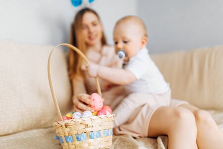 Mother and baby with rabbit ears sitting on the sofa with Easter eggs in a basket, parents and...