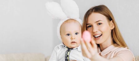 Mom and baby with rabbit ears, with Easter eggs in their hands, sitting on the sofa, parents and children play indoors. Family celebrates Easterの写真素材