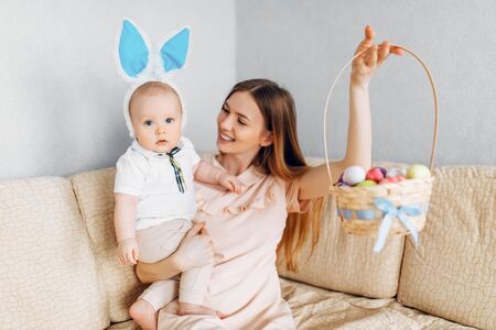 Happy Easter. A mother and a small baby, with rabbit ears on Easter day, with Easter painted eggs, have fun, play and cuddle, sitting on the sofa. Family celebrates Easterの写真素材