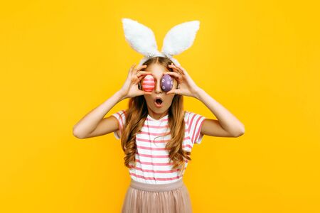 Shocked surprised beautiful girl on her head with rabbit ears and colorful Easter eggs in front of her eyes, on a yellow background. Symbol of Easter and spring.の写真素材