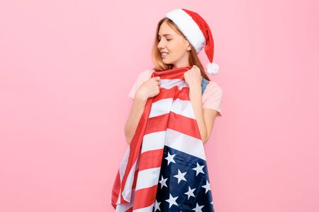 A smiling young stylish woman, wearing a Santa hat, holds the US flag on an isolated pink background. The Concept Of Christmasの写真素材