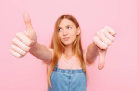 A young attractive stylish woman makes a good-bad sign. Undecided between Yes or no, on an isolated pink backgroundの写真素材