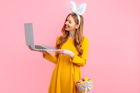 happy Easter. Happy young woman in the ears of an Easter Bunny holding a basket of colorful Easter eggs using a laptop on an isolated pink backgroundの写真素材