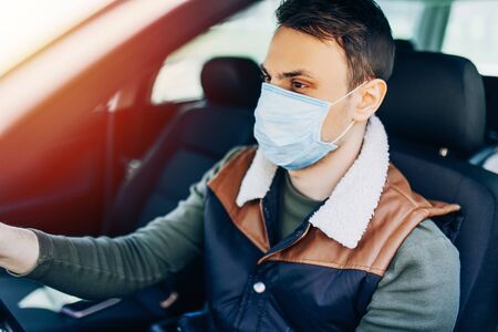 Beautiful young masked man sitting in a car, protective mask against coronavirus, driver on a city street during a coronavirus outbreak, covid-19の写真素材