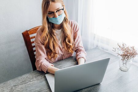 A young businesswoman works from home, sitting at a table with a laptop, wearing a protective mask. The concept of quarantine, coronavirusの写真素材