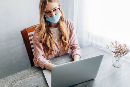 A young businesswoman works from home, sitting at a table with a laptop, wearing a protective mask. The concept of quarantine, coronavirusの素材 [FY310145417966]