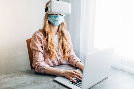 A young woman in a protective mask and virtual reality glasses, working at home during quarantine with a laptop. Quarantine, coronavirusの写真素材
