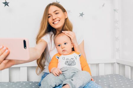 A happy mother and a small baby relax while sitting on the bed and take a selfie on their mobile...