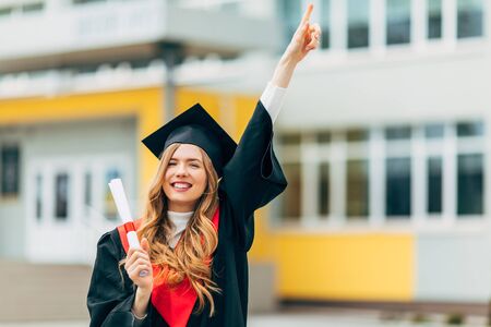 Happy beautiful girl student, in a master's dress, holding a diploma from the University. Concept of the graduation ceremonyの写真素材
